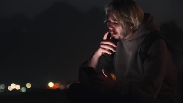 Uomo biondo con cappuccio beige seduto sulla spiaggia notturna a fumare una sigaretta. Mid shot - Filmati, video