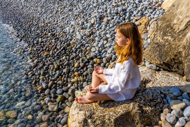 Όμορφο κοριτσάκι κάθεται σε ένα βράχο δίπλα στη θάλασσα και διαλογίζεται. Υψηλής ποιότητας φωτογραφία - Φωτογραφία, εικόνα