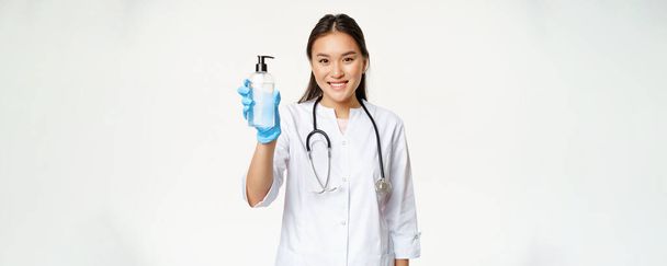 Lächelnder asiatischer Arzt mit Händedesinfektionsmittel, Flasche Antiseptikum, sterilen Gummihandschuhen und Klinikuniform, weißer Hintergrund. - Foto, Bild