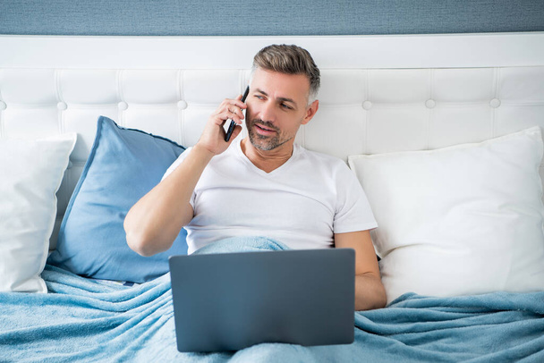 mature man talk on phone and working on laptop in bed. - Fotó, kép