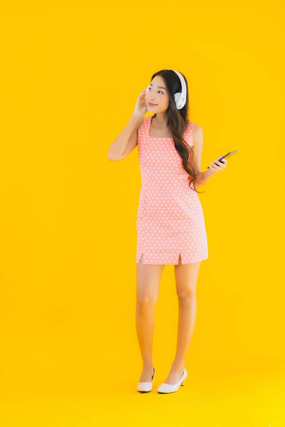 Güzel bir Asyalı kadın portresi. Sarı izole edilmiş arka planda akıllı cep telefonu ve kulaklıkla müzik dinliyor. - Fotoğraf, Görsel