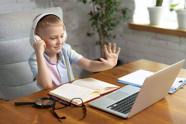 Greeting teacher. Little boy, school age kid sitting at home and doing homework, using laptop at home interior. On-line education, childhood, people, homework and school concept. Looks happy, calm - Фото, изображение