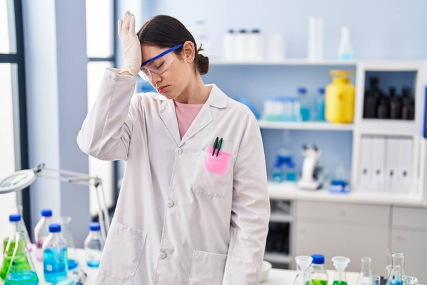 Young brunette woman working at scientist laboratory surprised with hand on head for mistake, remember error. forgot, bad memory concept.  - Foto, afbeelding