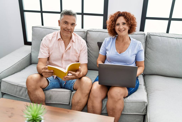 Couple homme / femme d'âge moyen utilisant un ordinateur portable et un livre de lecture assis sur un canapé à la maison - Photo, image