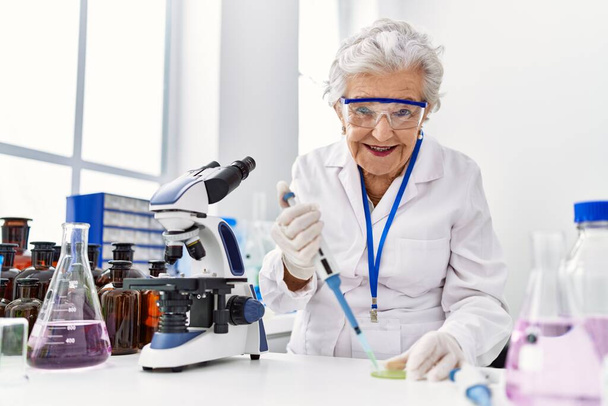 Mujer mayor de cabello gris usando uniforme científico usando pipeta en el laboratorio - Foto, imagen
