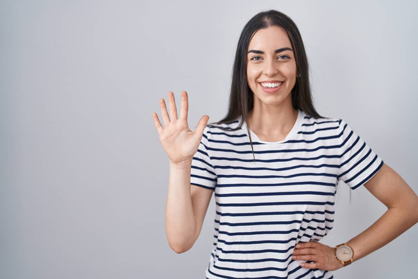 Young brunette woman wearing striped t shirt showing and pointing up with fingers number five while smiling confident and happy.  - Φωτογραφία, εικόνα