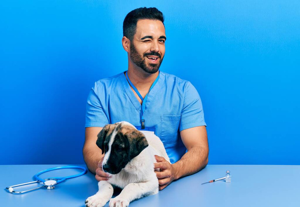Красивий ветеринарний чоловік з бородою перевіряє здоров'я собаки, підморгнувши, дивлячись на камеру з сексуальним виразом, веселим і щасливим обличчям
.  - Фото, зображення