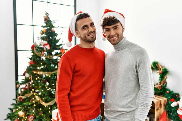 Young gay couple standing by christmas tree wearing hat looking away to side with smile on face, natural expression. laughing confident.  - Photo, Image