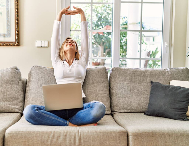 Mujer rubia joven estirando los brazos utilizando el ordenador portátil en casa. - Foto, imagen