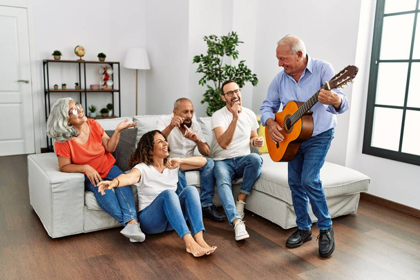 Grupo de amigos de mediana edad que tienen fiesta tocando la guitarra clásica sentados en el sofá en casa. - Foto, imagen