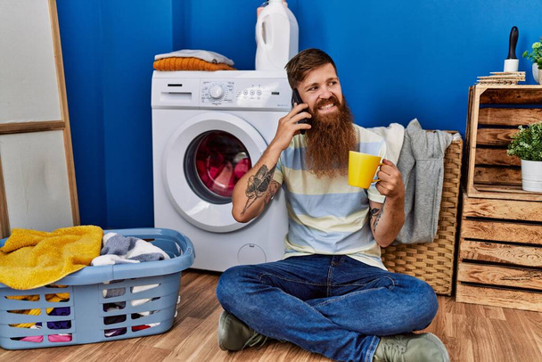 Молодий рудий чоловік розмовляє по смартфону і п'є каву в пральні
 - Фото, зображення