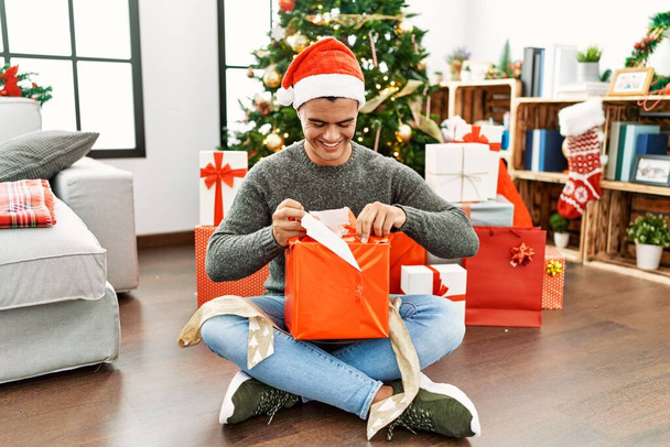 Jeune homme hispanique déballer cadeau assis par arbre de Noël à la maison - Photo, image
