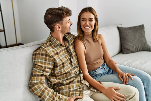 Νεαρό ζευγάρι ισπανόφωνων χαμογελά ευτυχισμένο και αγκαλιάζει κάθεται στον καναπέ στο σπίτι. - Φωτογραφία, εικόνα