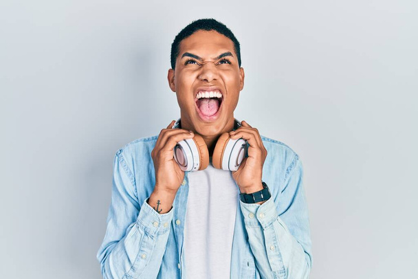 Młody Afroamerykanin słuchający muzyki za pomocą słuchawek wściekły i szalony krzyczy sfrustrowany i wściekły, krzycząc ze złości patrząc w górę.  - Zdjęcie, obraz