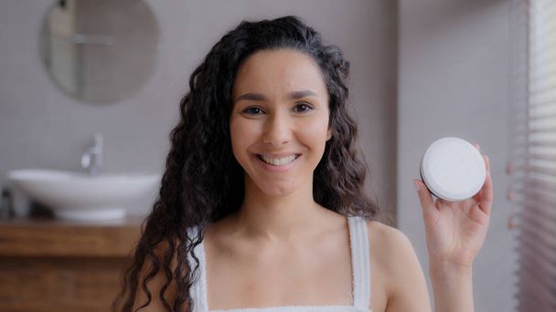 Крупним планом молода приваблива щаслива іспаномовна жінка, що стоїть у ванній кімнаті, дивлячись на камеру усміхнений продукт по догляду за шкірою, вказуючи на зволожуючий крем для банок, сприяє доведеній концепції ліків від шкіри
 - Фото, зображення