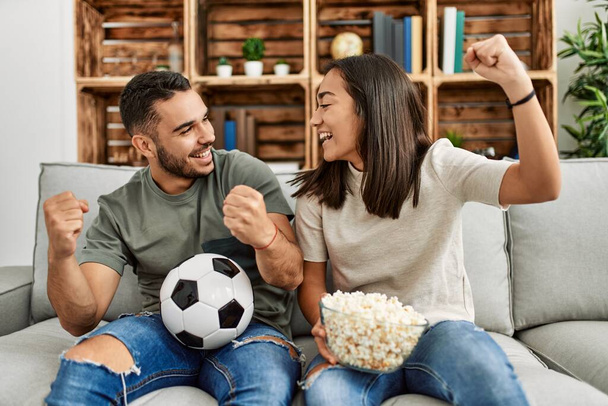 Joven pareja latina viendo un partido de fútbol comiendo palomitas en casa. - Foto, imagen