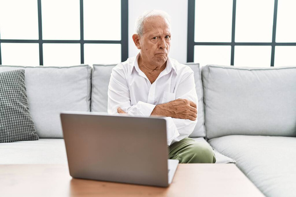 Vanhempi mies käyttää kannettavaa kotona istuu sohvalla skeptinen ja hermostunut, paheksuva ilme kasvot ristissä kädet. negatiivinen henkilö.  - Valokuva, kuva