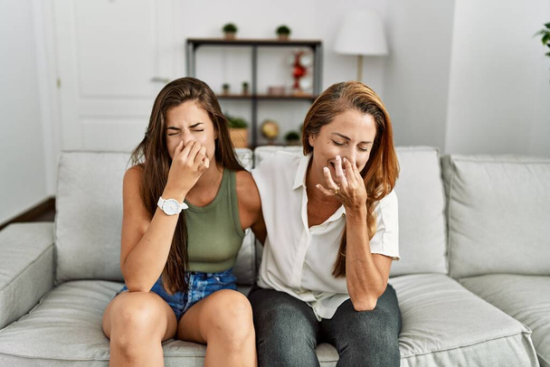 Mother and daughter together sitting on the sofa at home smelling something stinky and disgusting, intolerable smell, holding breath with fingers on nose. bad smell  - Photo, Image