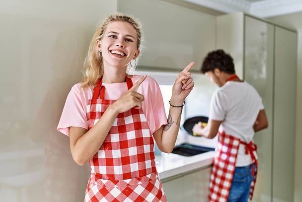 Young caucasian woman wearing apron and husband doing housework washing dishes smiling and looking at the camera pointing with two hands and fingers to the side.  - Φωτογραφία, εικόνα