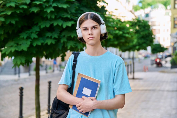 Serious teenage male student with textbooks looking at camera outdoor. Handsome guy 17, 18 years old with backpack headphones on city street. Youth, education, lifestyle, people concept - Photo, Image