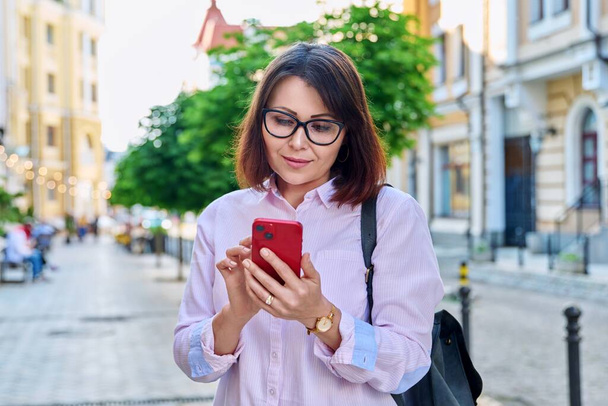 Middle aged smiling woman looking into smartphone on city street. Beautiful mature female using mobile online applications. Technology, business, lifestyle, urban style, 40s age people concept - Zdjęcie, obraz