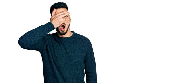 Young hispanic man with beard wearing casual winter sweater peeking in shock covering face and eyes with hand, looking through fingers afraid  - Foto, Imagen