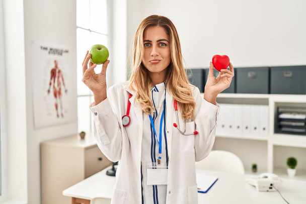 Young blonde doctor woman holding heart and green apple depressed and worry for distress, crying angry and afraid. sad expression.  - Photo, Image