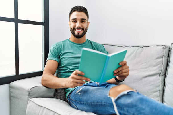 Young arab man reading book lying on sofa at home - Foto, immagini