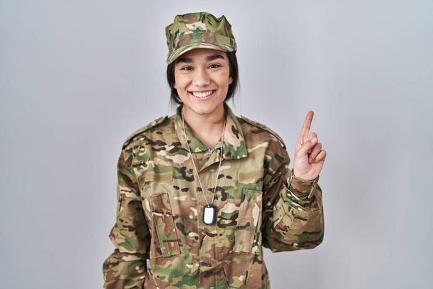 Mladý jižní asijské žena na sobě maskování armádní uniforma ukazující a ukazující nahoru s prstem číslo jedna při úsměvu sebevědomý a šťastný.  - Fotografie, Obrázek