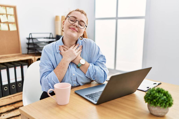 オフィスで働いている若い赤毛の女性は、目を閉じて胸の上で手で笑ってコンピュータのラップトップを使用して顔に感謝のジェスチャー。健康の概念.  - 写真・画像