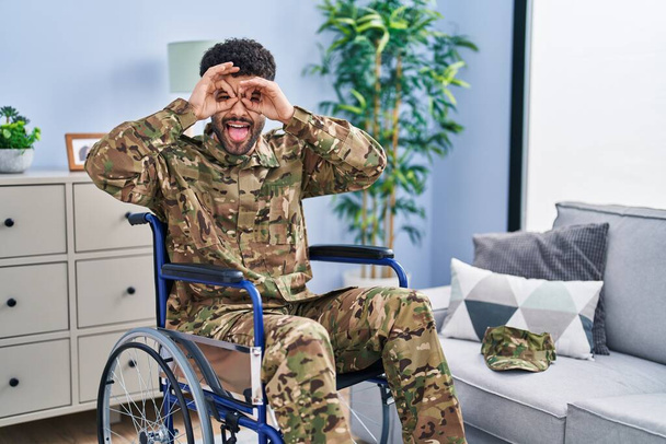 Homem árabe vestindo uniforme do exército de camuflagem sentado em cadeira de rodas fazendo gesto ok como binóculos furando a língua para fora, olhos olhando através dos dedos. expressão louca.  - Foto, Imagem
