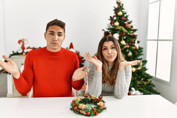Jovem casal hispânico sentado à mesa no Natal expressão indolente e confuso com braços e mãos levantadas. conceito de dúvida.  - Foto, Imagem