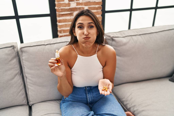 Young hispanic woman holding pills and cbd oil puffing cheeks with funny face. mouth inflated with air, catching air.  - Photo, Image