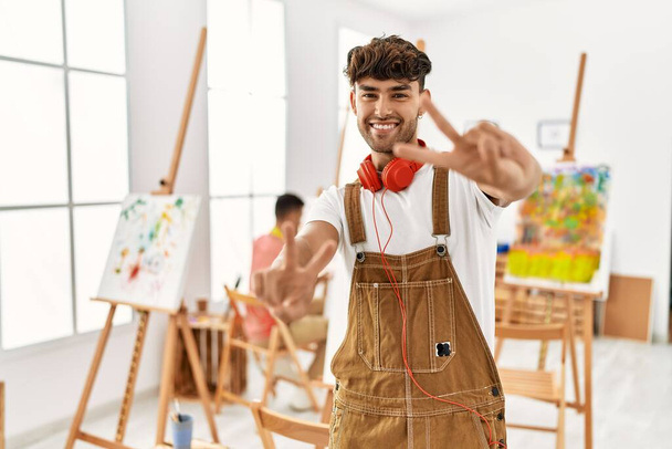 Mladý hispánec v uměleckém studiu se usmívá a dívá se na kameru ukazující prsty ukazující znamení vítězství. číslo dvě.  - Fotografie, Obrázek