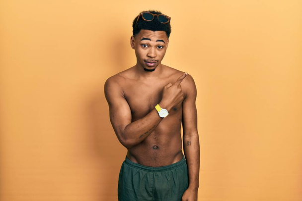 Junger afrikanisch-amerikanischer Mann mit Badebekleidung und Schwimmbrille, der mit dem Finger zur Seite zeigt und Werbung zeigt, ernstes und ruhiges Gesicht  - Foto, Bild