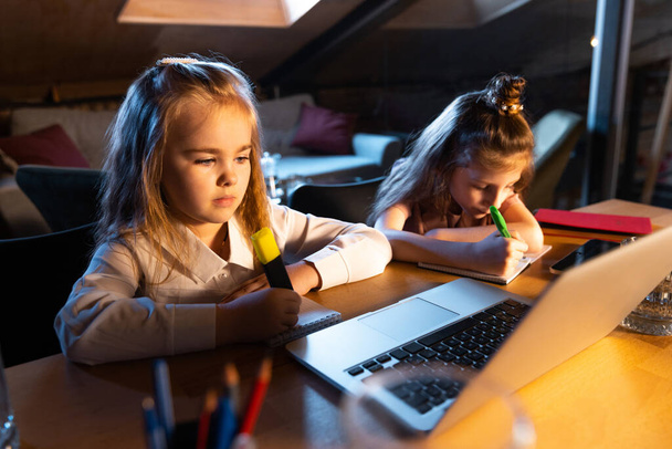 Studying with friend. Two little cute kids, sisters sitting at home and doing homework at home interior at evening time. Online education, childhood, family, remote learning and school concept. - Φωτογραφία, εικόνα