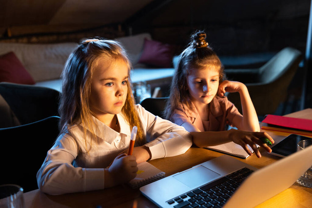 Leisure activities. Live portrait of cute kids, sisters sitting at home play computer games, chat at evening time. Online education, childhood, family, remote learning and school concept. - Фото, изображение