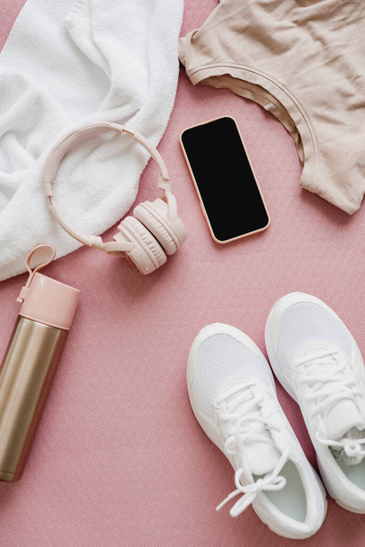 Flat lay of female sports clothing, smartphone, headphones, and a bottle of water on pink background. Training concept. - Zdjęcie, obraz