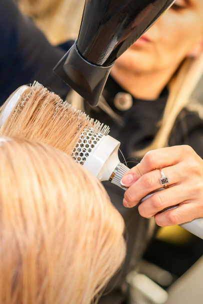 Séchage cheveux raides blonds avec sèche-cheveux noirs et brosse ronde blanche dans le salon de coiffure, gros plan - Photo, image