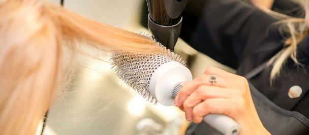 Сушка прямого світлого волосся з чорним феном і білою круглою щіткою в перукарні, крупним планом
 - Фото, зображення