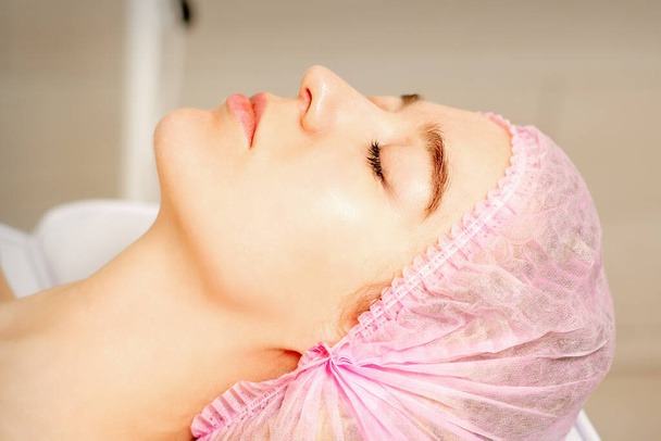 Vista lateral de una joven mujer caucásica con una gorra médica acostada con los ojos cerrados esperando el procedimiento cosmético en un salón de belleza - Foto, Imagen