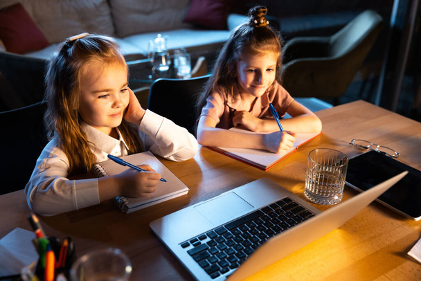 Studying with friend. Two little cute kids, sisters sitting at home and doing homework at home interior at evening time. Online education, childhood, family, remote learning and school concept. - Photo, image
