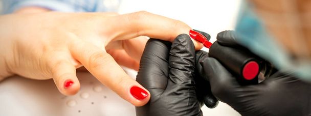 Manicure varnish painting. Close-up of a manicure master wearing rubber black gloves applying red varnish on a female fingernail in the beauty salon - Photo, Image