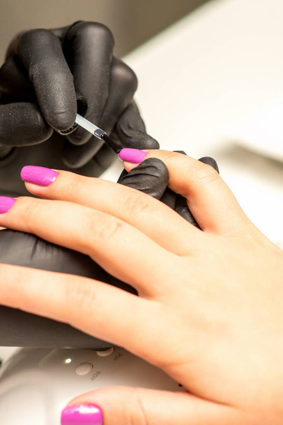 Professionele manicure. Een manicure schildert de vrouwelijke nagels van een cliënt met paarse nagellak in een schoonheidssalon, close-up. Begrip schoonheidssector - Foto, afbeelding