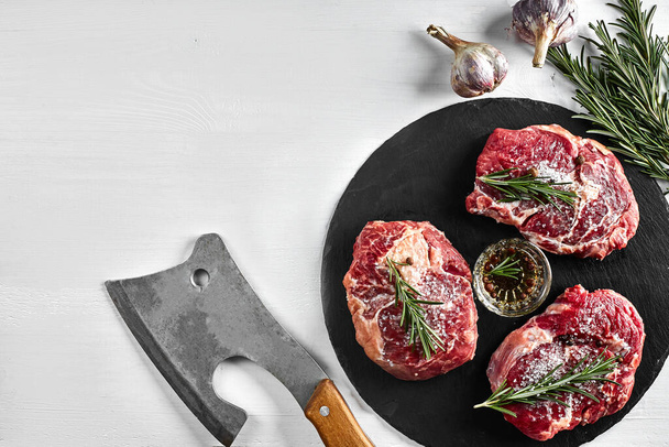 Fresh raw beef steaks with herbs, garlic, olive oil, pepper, salt and rosemary on black board: Tenderloin, Striploin, Rib Eye and a kitchen ax on white background. Top view. Copy space. Still life - Foto, Imagem