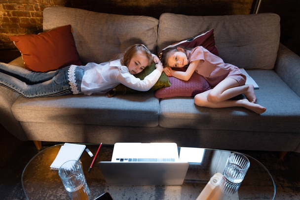 Look tired, sleeping. Cute little girls, kids having fun, watching cartoons on laptop at home at evening time. Education, childhood, family, remote learning and emotions concept. - Фото, зображення