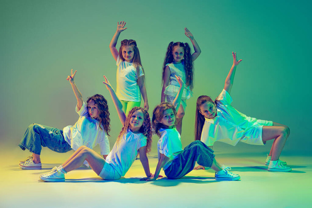 Future contemp dancers. Group of children, little girls in sportive casual style clothes dancing in choreography class isolated on green background in neon light. Concept of music, fashion, art - 写真・画像