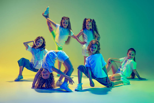 Hip-hop dance, street style. Group of children, little girls in sportive casual style clothes dancing in choreography class isolated on green background in neon light. Concept of music, fashion, art - Fotó, kép