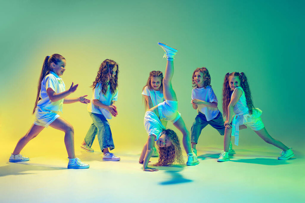 Happy children dancing. Group of children, little girls in sportive casual style clothes dancing in choreography class isolated on green background in yellow neon light. Concept of music, fashion, art - Foto, Bild