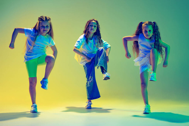 Hip-hop dance, street style. Group of children, little girls in sportive casual style clothes dancing in choreography class isolated on green background in neon light. Concept of music, fashion, art - Foto, Imagen